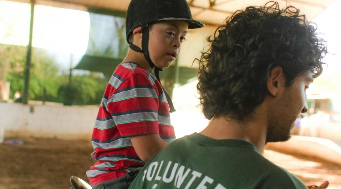 Male Equine Therapy intern working with children in Argentina leads a horse ridden by a child during a therapy session.