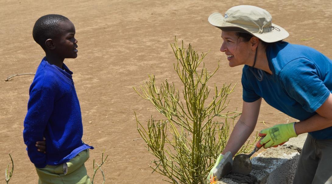 A teenage volunteer doing construction work in Tanzania builds a wall while chatting to a young child.  